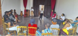 Participants assis en cercle sur des chaises dorées dans une salle aux rideaux rouges, écoutant un intervenant debout au centre, Musée national du Cameroun, mars 2025.