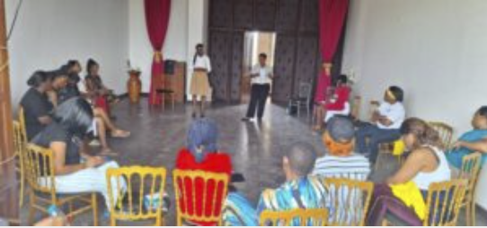 Participants assis en cercle sur des chaises dorées dans une salle aux rideaux rouges, écoutant un intervenant debout au centre, Musée national du Cameroun, mars 2025.